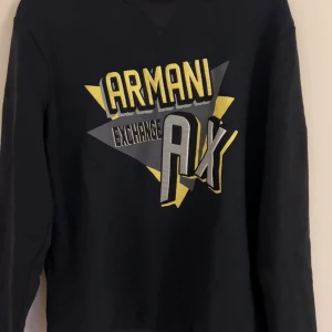 Svart Armani Exchange sweatshirt - Svart sweatshirt från Armani Exchange med stort grafiskt tryck i gult, grått och vitt framtill. Tröjan har lång ärm och klassisk rund hals. Perfekt för dig som gillar streetstyle och vill sticka ut med en bold logga.