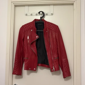 Röd skinnjacka från Zara Woman - Snygg röd skinnjacka från Zara Woman med quiltade detaljer på axlar och ärmar, dragkedjor och spännen i silver. Jackan har en klassisk bikerstil med flera fickor och coola bältesdetaljer. Perfekt för dig som vill sticka ut med en edgy look.