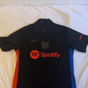 FC Barcelona Nike fotbollströja svart - Snygg svart FC Barcelona fotbollströja från Nike med röda och blå sidopaneler. Tröjan har korta ärmar, klubbmärke och Nike-logga på bröstet samt Spotify-tryck. Materialet är lätt och andas, perfekt för match eller träning.