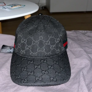 Svart keps från Gucci med GG-mönster - Säljer en svart keps från Gucci med klassiskt GG-monogram över hela kepsen och gröna/röda ränder på sidorna. Justerbart spänne bak i svart skinn och detaljer i metall. Kepsen är tillverkad i polyester och bomull, perfekt för dig som vill sticka ut med en lyxig accessoar.