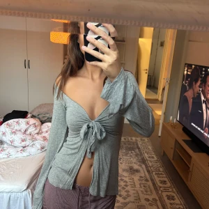 Grå omlottopp med knyt och v-ringning - Säljer en grå omlottopp med djup v-ringning och knyt framtill. Toppen är offshoulder med långa, lite utsvängda ärmar och croppad passform framtill. Mjuk och stretchig i bomullsmix, perfekt för en chill och trendig look.