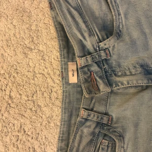 Low waist Ljusblå ripped jeans från Nelly - Säljer ett par ljusblå jeans från Nelly med slitna detaljer på båda benen och cool rosa söm på bakfickan. Klassisk femficksmodell med bälteshällor och raka ben. Perfekta för en avslappnad och trendig look.dem är uppsydda i benen men det går utmärkt att bara sprätta upp igen om så önskas