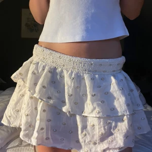 Vit volangkjol med guldiga detaljer - Supersöt vit kortkjol med volanger och små guldiga broderade prickar. Kjolen har bred smock i midjan för skön passform och är gjord i ett lätt, luftigt material. Perfekt för varma dagar när du vill känna dig extra fin.