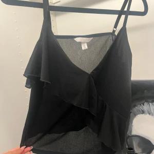 Svart volangig linnetopp H&M - Svart linnetopp från H&M med smala axelband och v-ringning. Toppen har en snygg volangdetalj framtill som ger en cool och trendig look. Tunt och lätt material som passar perfekt till varma dagar eller lager på lager.