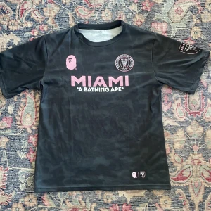 Inter Miami x BAPE fotbollströja svart - Svart fotbollströja från Inter Miami i samarbete med A Bathing Ape. Tröjan har rosa tryck med MIAMI-logga och BAPE-apan på bröstet, klubbmärke på bröstet och ärmen samt diskret camouflage-mönster i tyget. Tillverkad i lätt och sval polyester, perfekt för match eller streetwear.