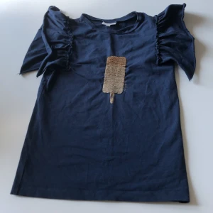 Mörkblå topp med volangärm Pomp de Lux - Supersöt mörkblå topp från Pomp de Lux med korta volangärmar och ett glittrigt paljettmotiv av en glass på bröstet. Toppen är i mjuk bomull och har en rak passform. Perfekt för dig som gillar unika detaljer och vill sticka ut lite extra.