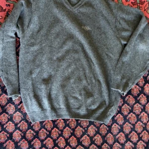 Grå v-ringad ulltröja HOL - Mysig grå v-ringad tröja från HOL i mjuk ullmix. Klassisk passform med ribbade muddar vid ärmslut och nederkant. Broderad logga på bröstet ger en clean och stilren look. Perfekt för lager-på-lager under kyliga dagar.