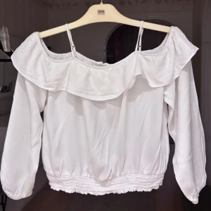 Vit offshoulder blus med volang - Supersöt vit blus med offshoulder och smala axelband. Volang upptill och smockad nederkant samt ärmslut ger en romantisk vibe. Långärmad och i mjukt, tunt tyg som känns luftigt och perfekt för varmare dagar.