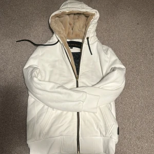 Moose knuckles Bunny jacka  - Säljer en snygg vit teddyjacka från Moose Knuckles med guldfärgade detaljer på ärmen och dragkedja. Jackan har en mysig beige fuskpäls i huvan och svart quiltat innerfoder. Perfekt för kalla dagar och riktigt stilren look. Kan även passa M