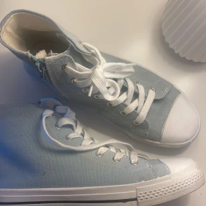 Ljusblå höga sneakers med dragkedja - Snygga ljusblå höga sneakers i canvas med vita snören och tåhätta. Skorna har dragkedja på sidan för enkel påtagning och klassisk vit sula med svart rand. Perfekta för dig som gillar en clean och fräsch stil.