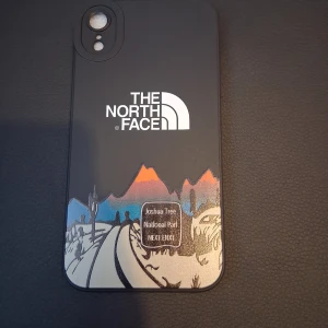 The North Face mobilskal till iPhone xr - Snyggt svart mobilskal från The North Face med coolt motiv av Joshua Tree National Park och färgglada berg i orange, blått och beige. Skalet har en stilren design med vit logga och illustration av landskap.
