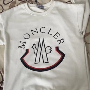 Vit sweatshirt från Moncler - Snygg vit sweatshirt från Moncler med stort tryck på bröstet i svart och rött. Klassisk rund halsringning och långa ärmar. Perfekt för dig som gillar stilrena och exklusiva plagg med tydlig logga. Mjuk och skön bomullskänsla.