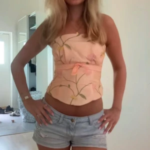 Persikofärgad blommig korsettopp - Superfin korsettopp i persikofärg med blommigt mönster och en söt rosett framtill. Toppen är axelbandslös och har en figurnära passform som framhäver midjan. Perfekt för varma dagar när du vill sticka ut lite extra.