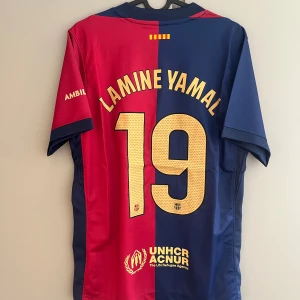 FC Barcelona Lamine Yamal tröja S - Säljer en officiell FC Barcelona fotbollströja från Nike med Lamine Yamal och nummer 19 på ryggen. Tröjan har klassisk röd och blå färgdelning, klubbmärke på bröstet, guldfärgade detaljer och tryck, samt korta ärmar. Materialet är lätt och andas, perfekt för match eller träning.