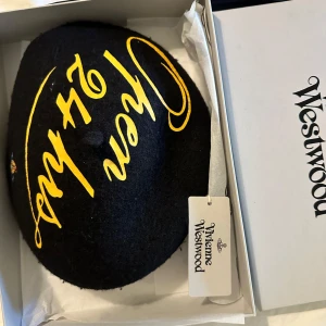 Svart basker från Vivienne Westwood - Svart ullbasker från Vivienne Westwood med gul broderad text och ikonisk orb-detalj i blått och beige. Klassisk rund form med liten topp och unik design som sticker ut. Perfekt accessoar för dig som vill ha något extra.
