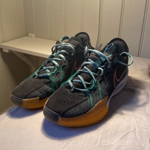 Nike sneakers med turkosa detaljer - Snygga svarta Nike GT CUT 3 sneakers med gröns snören och detaljer, orange swoosh och gul sula. Ovanför på skon är materialet rätt slitet med tyget som har lossnat lite, undersidan på skon är däremot nästan knappt påverkad av spel och har varit bra för mig. 44 i stl men passar säkert en del 43.5