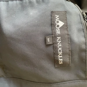 Svart dunjacka Moose Knuckles S - Säljer en svart dunjacka från Moose Knuckles i storlek S. Jackan har en fluffig huva med ljus päls, flera dragkedjor och snygga detaljer på ärmarna. Perfekt för kalla dagar och riktigt stilren look.