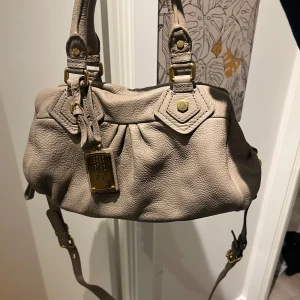 Beige skinnväska Marc by Marc Jacobs - Snygg beige handväska från Marc by Marc Jacobs i mjukt skinn med guldfärgade metalldetaljer. Väskan har både handtag och justerbar axelrem samt dragkedja upptill. Klassisk form med fina veck och dekorativa nitar.