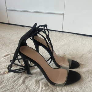 Svarta klackar med snörning och transparent rem - Stilrena svarta klackar med höga smala klackar, snörning runt vristen och transparent rem över tårna. Skorna har en rund tå och ovandel i mocka-look. Perfekta för dig som vill ha en trendig och minimalistisk look. Från PLT storlek 37