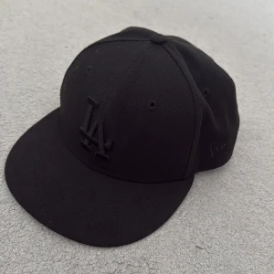 Svart LA-keps New Era 59FIFTY - Säljer en svart New Era 59FIFTY keps med LA-logga framtill i svart brodyr. Klassisk flat peak och strukturerad passform. Kepsen är tillverkad i polyester och bomull, perfekt för dig som gillar streetstyle och sportig look.