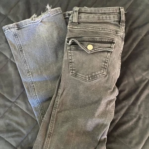 Grå jeansbyxor XXS med fickdetalj - Säljer ett par grå jeansbyxor  i storlek XXS. Byxorna har en snygg ficka med lock och knapp bak samt rå kant nertill på ena benet för en cool look. Perfekta för dig som gillar en avslappnad men trendig stil.
