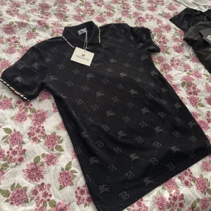 Burberry piké - Svart pikétröja från Burberry med diskret burberry logga och ryttare tryckt över hela plagget. Klassisk krage med tunna ränder och korta ärmar med randiga muddar. Tillverkad i mjuk bomull med lite elastan för extra komfort. Perfekt för en clean och stilren look. Står L men är M/S‼️‼️❗️❗️