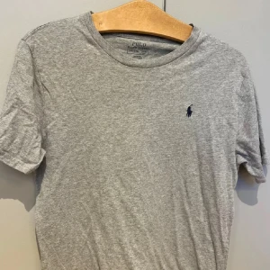 Grå t-shirt från Polo Ralph Lauren - Säljer en grå t-shirt från Polo Ralph Lauren med den klassiska häst loggan på bröstet, tishan är i grymt skick knappast använd extremt fåtal gånger. Storlek 160 (XS) 14-16 år. Inga slitningar, KÖP!