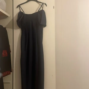 Mörkblå långklänning med puffärm - Snygg mörkblå långklänning från NA-KD med tunna axelband och puffiga offshoulder-ärmar. Klänningen har en slits på sidan och är gjord i ett glansigt, satinliknande material som ger en lyxig känsla. Perfekt för dig som vill sticka ut med en elegant och trendig look.