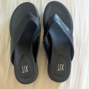 Svarta sandaler från XIT med platåsula - Stilrena svarta sandaler från XIT med breda remmar och platåsula. Skorna har en minimalistisk design med rund tå och är tillverkade i syntetmaterial. Perfekta för sommardagar när du vill ha en clean och enkel look. Endast använda en gång säljs pga används inte men super snygga! Köparen står för frakt:)