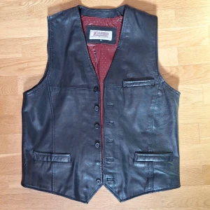 Svart skinnväst - Svart äkta skinnväst från Jofama med klassisk v-ringning och knäppning framtill. Västen är vinröd på insidan, har tre fickor fram och ett justerbart bälte bak. En knapp saknas och det finns några små igensydda hål på insidan men i övrigt väldigt bra skick.