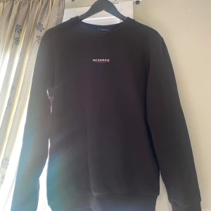 Svart sweatshirt från McKenzie - En stilren svart sweatshirt från McKenzie i storlek M. Tröjan har rund halsringning, lång ärm och en enkel vit logga broderad på bröstet. Tillverkad i mjuk bomullsblandning som är skön att ha på sig hela dagen.