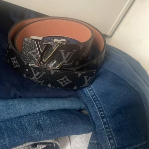  svart skärp med monogram - Säljer ett svart bälte med klassiskt monogrammönster och stor spänne i silver. Bältet är gjort i skinn med en insida i ljusbrun ton. Perfekt accessoar för att lyfta din outfit.