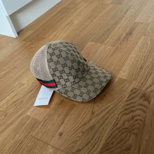 GG keps - Snygg beige keps med klassiskt GG-mönster över hela kepsen och ikoniska gröna och röda ränder på sidan. Kepsen har böjd skärm och mesh baktill för extra ventilation. Perfekt accessoar för att lyfta din outfit.