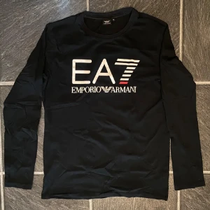 Svart långärmad tröja EA7 Armani - Svart långärmad tröja från Emporio Armani EA7 med stor vit och röd logga framtill. Klassisk rund halsringning och stilren design som passar perfekt till jeans eller joggers. Mjuk bomullskänsla och snygg passform.