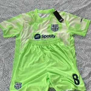 FC Barcelona Pedri fotbollströja set - Snygg FC Barcelona fotbollströja med Pedri och nummer 8 på ryggen. Tröjan är ljusgrön med vita detaljer och har klubbmärke, Spotify-logga och UNHCR-tryck. Matchande shorts med nummer 8 och klubbmärke. Tillverkad i lätt och ventilerande material.