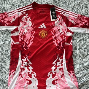 Manchester United x Adidas röd matchtröja - Manchester United fotbollströja från Adidas med unikt röd och rosa grafiskt mönster, vita detaljer och klubbmärke broderat på bröstet. Tröjan är kortärmad och tillverkad i lätt funktionsmaterial (AEROREADY) som andas. Perfekt för dig som vill sticka ut på planen.