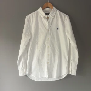 Vit skjorta från Ralph Lauren S - Klassisk vit skjorta från Ralph Lauren i bomull med button down-krage och broderad blå logga på bröstet. Långärmad modell med knappar framtill och rundad nederkant. Perfekt för en clean och stilren look. Tröjan är helt ny hör av dig vid frågor och funderingar 