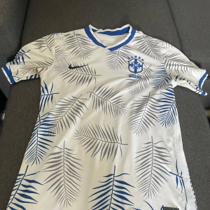 Vit Brasilien fotbollströja Nike - Snygg vit Brasilien fotbollströja från Nike med blå och grå palmbladsmönster över hela tröjan. Broderat CBF-emblem på bröstet och blå detaljer vid krage och ärmslut. Tillverkad i lätt och ventilerande material, perfekt för match eller träning.