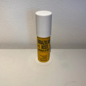 Brazilian Kiss Cupuaçu Lip Butter - Läppbalsam från Sol de Janeiro i en gul och vit tub med texten 'Brazilian Kiss Cupuaçu Lip Butter'. Innehåller cupuaçusmör som ger läpparna fukt och en mjuk känsla. Doftar sött och tropiskt. Perfekt att ha i väskan för återfuktade läppar hela dagen.