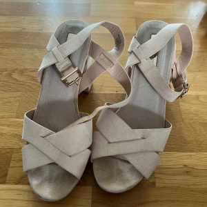 Beige sandaler med klack och platå - Snygga beige sandaler med breda korslagda remmar och justerbart spänne i guld. Skorna har öppen tå, klack och platåsula för extra höjd. Tillverkade i mockaliknande material som ger en mjuk känsla. Perfekta för dig som vill ha en stilren och trendig look.