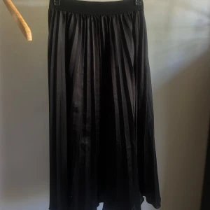 Svart plisserad långkjol Cherry Koko - Svart långkjol från Cherry Koko med plisserad design och bred resår i midjan. Kjolen har ett snyggt fall och passar perfekt till sneakers eller boots. Materialet känns lätt och flowy, vilket ger en chill och stilren vibe.