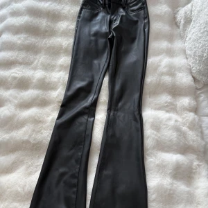 Svarta bootcut byxor i skinnlook Vero Moda - Snygga svarta byxor från Vero Moda i skinnimitation med bootcut-ben och hög midja. Klassisk femficksmodell med bälteshällor och dragkedja. Perfekta för dig som vill ha en edgy vibe och stilren look.