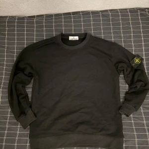 Svart sweatshirt från Stone Island - Svart sweatshirt från Stone Island med klassisk rund hals och ribbade muddar. Ikonisk logotyp-patch på vänster ärm som går att ta av. Tröjan är i mjuk bomull och har en clean, stilren look som passar perfekt till streetwear. Bara använt 2 gånger. Hör av er om ni har några frågor.