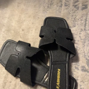 Svarta sandaler med fyrkantig tå - Stilrena svarta sandaler med fyrkantig tå och breda remmar i präglat syntetmaterial. Snyggt minimalistisk design med öppen tå och platt sula, perfekt för varma dagar. Texten 'FASHION' i guld på innersulan ger en trendig touch.