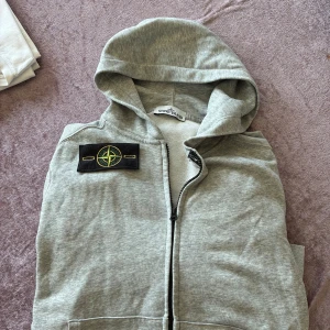 Grå hoodie från Stone Island M - Snygg grå hoodie från Stone Island med klassisk kompasspatch på ärmen. Tröjan har dragkedja framtill, huva och är tillverkad i mjuk bomull. Perfekt för dig som gillar streetwear och vill ha en stilren look med premiumkänsla.
