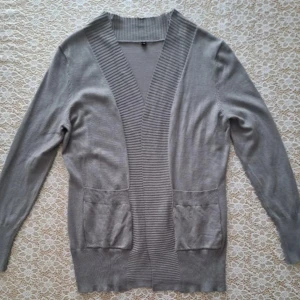 Ljusgrå kofta med fickor - En stilren ljusgrå kofta med djup ribbad kant framtill och två praktiska fickor. Koftan är långärmad och har en mjuk, tunn stickad känsla som gör den perfekt att slänga över en t-shirt eller topp. Enkel att matcha och passar till många olika looks.