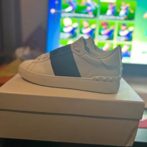 Valentino Garavani Open sneakers vit/blå - Snygga Valentino Garavani Open sneakers i vitt skinn med bred blå rem över mitten. Klassisk rund tå, vita snören och ikoniska nitar baktill på sulan. Perfekta för dig som vill ha en clean men lyxig look. Skriv vid intresse pris kan diskuteras