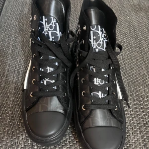 Dior B23 Storlek 44 - Exklusiva Dior high-top sneakers i svart och vitt med ikoniskt Dior-mönster. Skorna har snörning, rund tå och platt sula. Tillverkade i canvas och PVC med tydliga logodetaljer på sidan och hälen. Perfekt för dig som vill sticka ut med lyxig streetstyle.