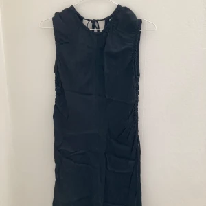 Svart ärmlös klänning med rynkade detaljer - Säljer en svart ärmlös klänning med snygga rynkade detaljer framtill och baktill. Klänningen är från Arket har knytband i midjan för justerbar passform och är tillverkad i ett mjukt, lätt material som ger ett elegant fall.