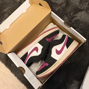 Nike Air Jordan 1 Low svart/vit/rosa - Säljer ett par Nike Air Jordan 1 Low i svart, vitt och rosa. Skorna har klassisk låg siluett med svart ovandel, vita paneler och rosa detaljer på swoosh och tå. Perfekta sneakers för dig som vill sticka ut med stil.